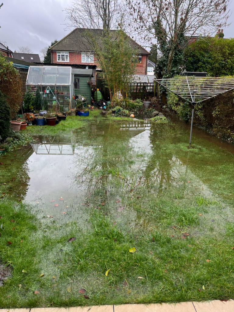 Flooded garden with standing water on grass and patio, greenhouse visible.