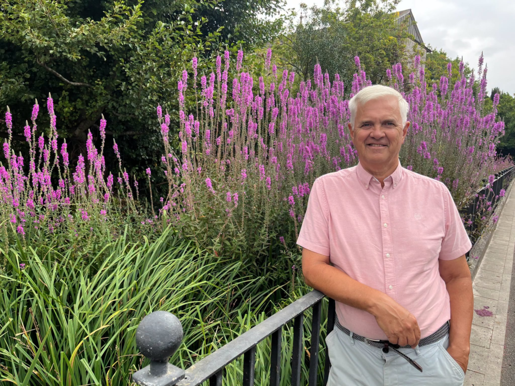 Man in pink shirt smiles by purple flowers, potentially related to app development trends.