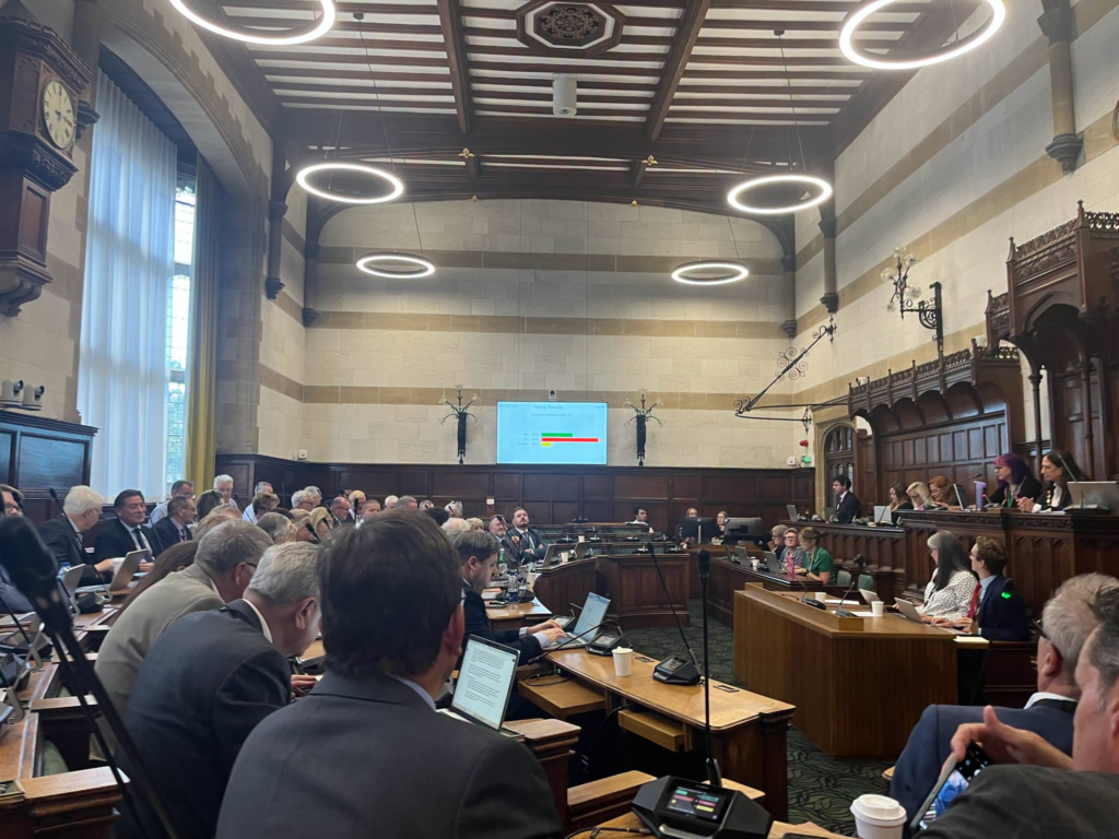 Meeting in a grand hall with a screen displaying "New Forest Parking Charges