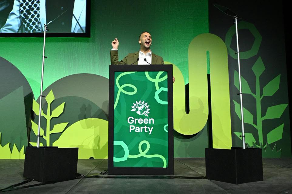 Green Party speaker at podium with "Green Party" logo on screen