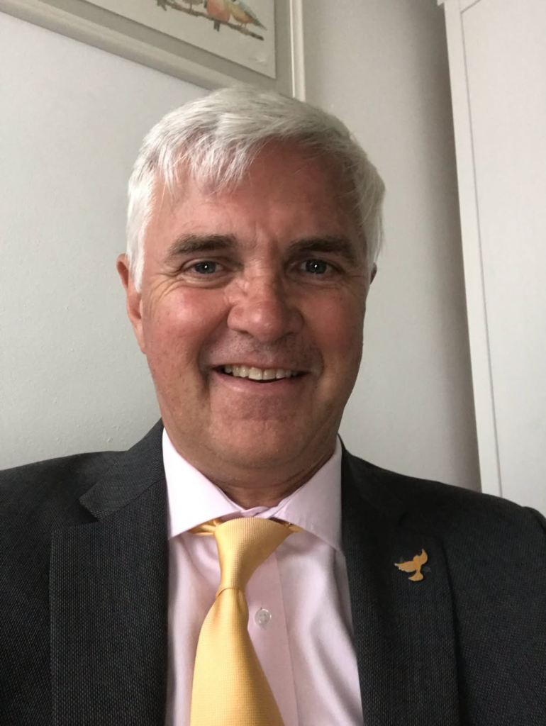 Smiling man in suit and yellow tie with bird pin, possibly discussing public services.