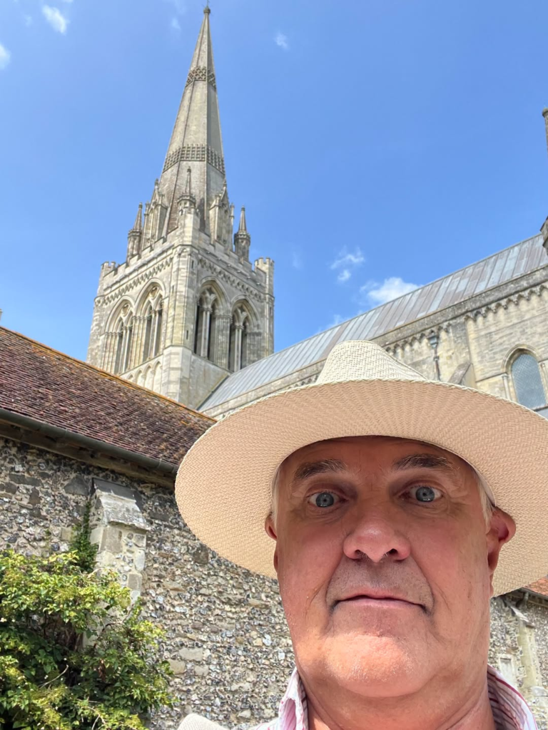 Man in straw hat takes selfie with historic cathedral spire behind him.