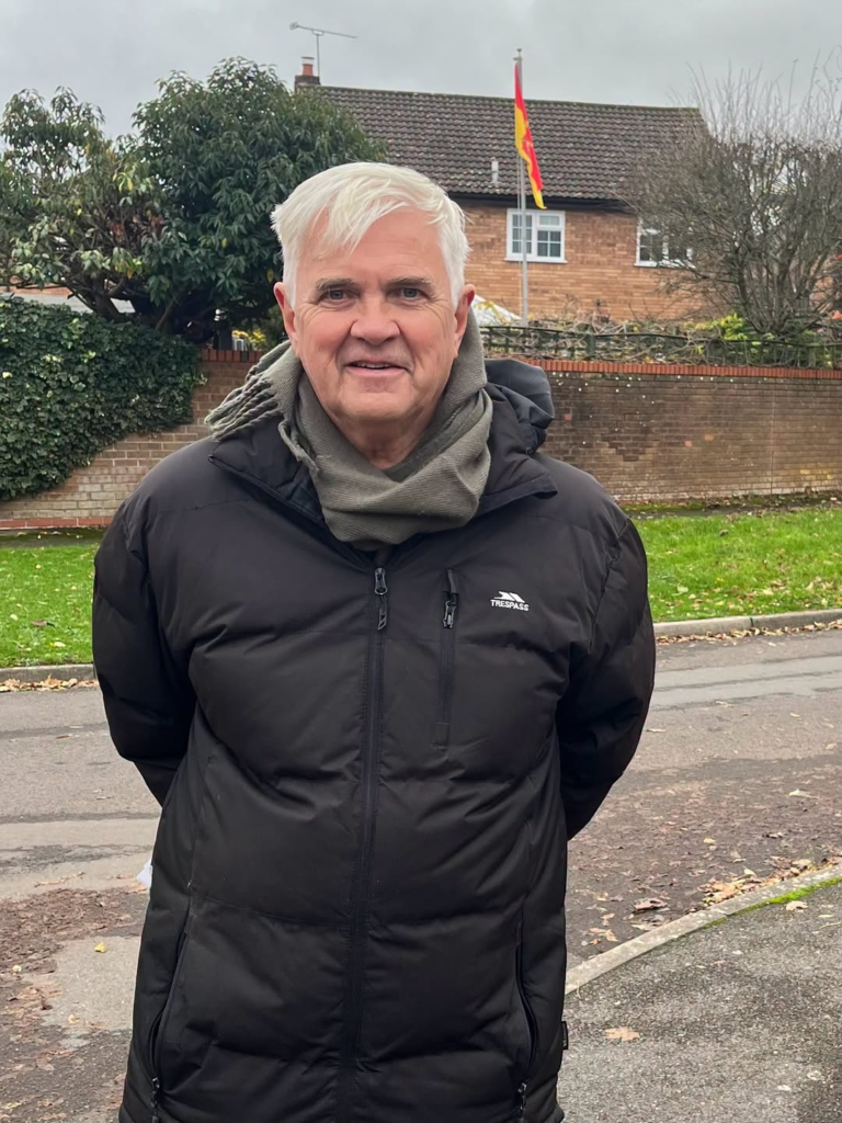 Man in puffer jacket and scarf smiles outdoors, house and flag in background
