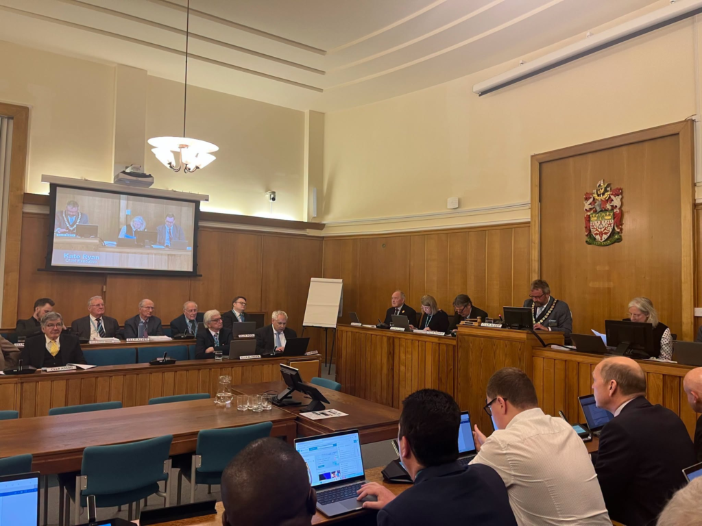Council members in meeting, discussing budget cuts or savings.