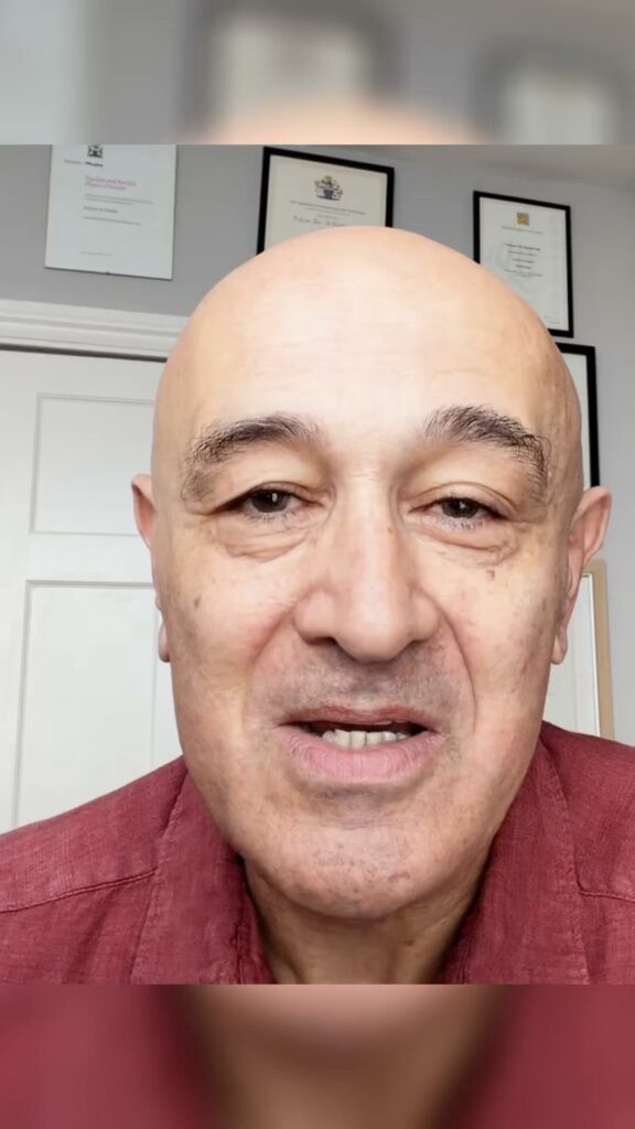 Bald man in red shirt, speaking, with framed certificates on wall