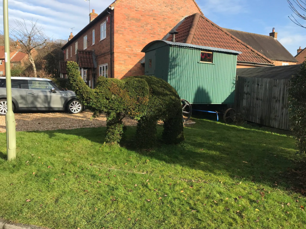 Elephant-shaped topiary on a lawn, with a green shepherd's hut and car in the background.