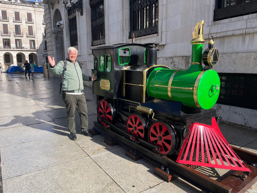 Councillor David Harrison waves next to a miniature green train in a city square.