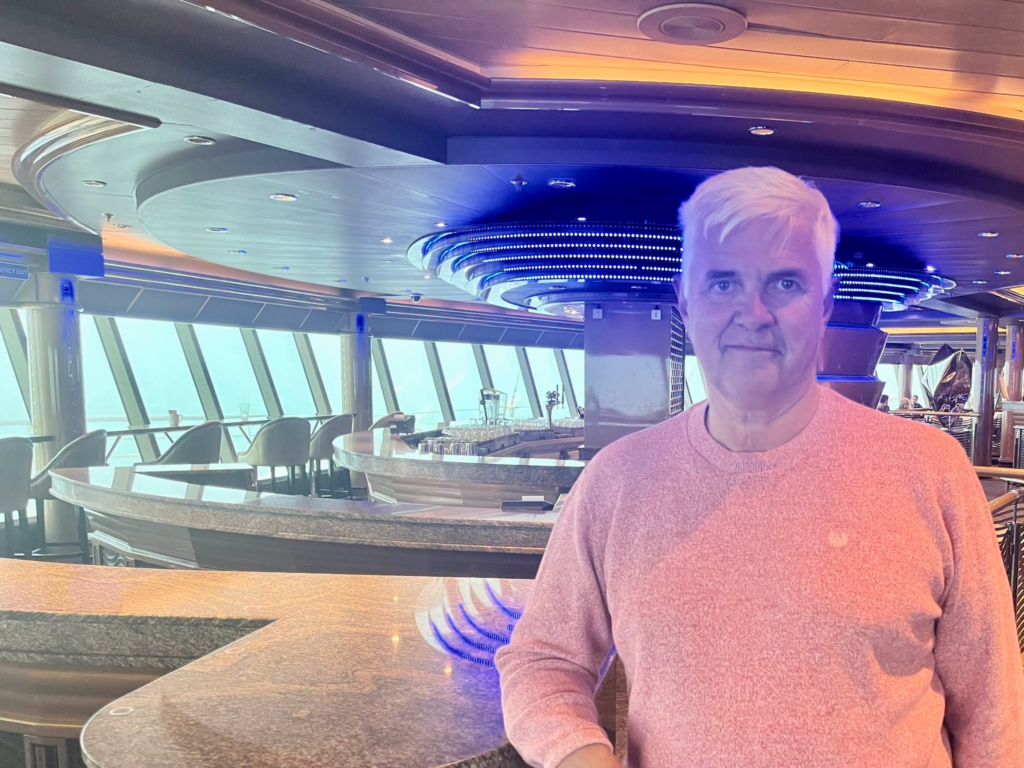 Man in pink sweater stands in a modern cruise ship lounge with ocean view.